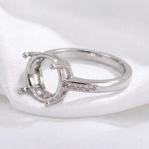 May include: A silver-toned metal ring setting featuring a circular prong head designed to hold a gemstone. The band is accented with a row of small, clear, round-cut stones embedded along the side of the shank, creating a polished finish.