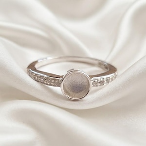 Könnte beinhalten: Ein silberfarbener Ring mit einer zentralen, kreisförmigen Fassung für einen Stein oder Cabochon. Die Ringschiene ist mit einer Reihe kleiner, klarer, funkelnder Steine besetzt, die beidseitig entlang der Schiene bis zur Mitte verlaufen.