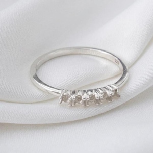Könnte beinhalten: Ein silberner Ring mit fünf Krappen zur Aufnahme von Edelsteinen. Der Ring ist vor einem weißen Hintergrund platziert, der seine polierte Oberfläche hervorhebt. Das Band ist schmal, mit einer kleinen Markierung innen.