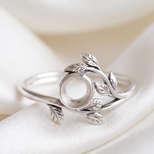 Op de afbeelding: Een zilverkleurige metalen ring met een ronde zetting voor een steen, versierd met een delicaat takmotief dat verschillende kleine, getextureerde blaadjes bevat die zich langs de ringband aan beide zijden van de centrale houder uitstrekken.