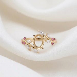 以下が含まれることがあります： 楕円形のセッティングと葉のようなデザインが特徴のゴールドリング。リングには、小さな白と赤の宝石が使用されています。白い背景にリングが置かれています。