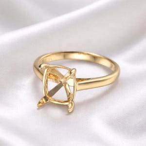 May include: A gold-colored ring setting with a square center for a gemstone. The ring has a smooth, polished band and prongs to hold the stone. The ring is displayed on a white, silky background.