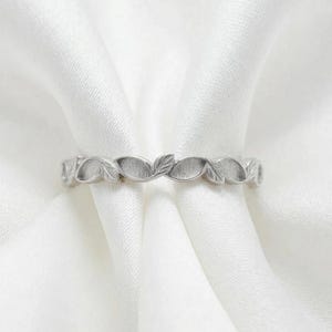 May include: A silver ring with a repeating leaf design. The ring is made of a shiny metal and features a series of stylized leaves that encircle the band. The leaves are evenly spaced and create a delicate, nature-inspired pattern.