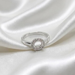 Può includere: Un anello d'argento con incastonatura rotonda, circondato da piccole pietre trasparenti. La fascia dell'anello è liscia e sottile, con la scritta "S925" visibile. L'anello è presentato su un tessuto bianco e serico.