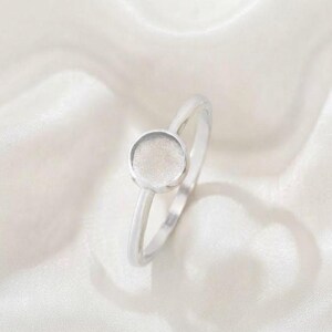 Könnte beinhalten: Ein silberfarbener Ring mit einer dünnen, glatten Ringschiene und einer kreisförmigen, flachen Fassung. Die Mitte des kreisförmigen Elements weist eine matte, texturierte Oberfläche auf, während die Schiene und der Rand glänzend poliert sind.