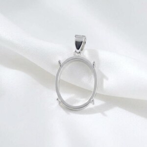 May include: A silver-colored oval pendant setting with a loop at the top for a chain. The setting has four prongs to hold a stone. The pendant is displayed on a white, textured fabric.
