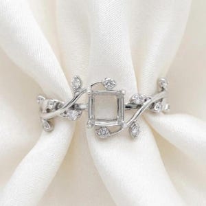 Könnte beinhalten: Ein silberfarbener Ring mit einer zentralen rechteckigen Fassung für einen Edelstein. Die Ringschiene ist mit einem rankenartigen Motiv gestaltet, in das kleine, klare, runde Steine in die Metallblätter eingearbeitet sind.