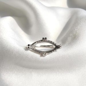 May include: A silver-colored ring setting with an oval shape, featuring small decorative elements and a row of tiny stones along the edges. The ring is set against a white, silky background.