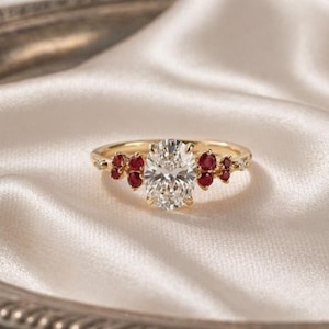 Könnte beinhalten: Ein goldener Ring mit einem oval geschliffenen Diamanten als Mittelstein und kleinen roten Edelsteinen auf beiden Seiten. Der Ring liegt auf einer weißen Satin-Oberfläche, was ein luxuriöses Gefühl erzeugt. Das Design des Rings ist elegant und auffällig.