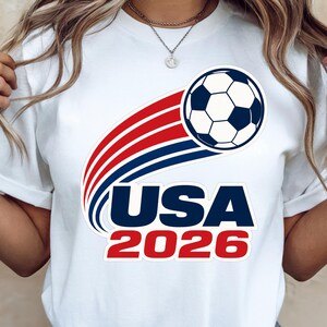 May include: White t-shirt featuring a soccer ball graphic with red and blue curved lines. The text "USA 2026" is printed below the soccer ball, in blue and red. The shirt is a casual style.