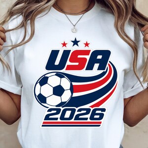 May include: White t-shirt with a graphic design featuring the text "USA 2026" above a soccer ball and red, white, and blue stripes. The shirt has a crew neck and short sleeves.