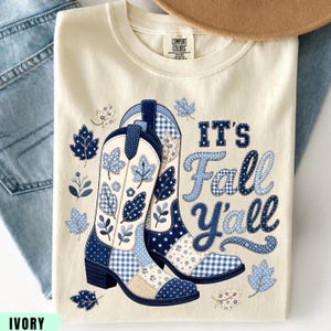 May include: An ivory t-shirt featuring a graphic of two cowboy boots decorated with blue and white patchwork patterns, including gingham, stars, and leaves. The text "IT'S Fall Y'all" is printed in a blue, stitched-style font alongside scattered leaf motifs.