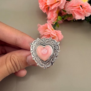 May include: A silver ring featuring a central heart-shaped pink shell cabochon. The setting is crafted with intricate metalwork, including a twisted rope border and a decorative scalloped frame with small circular cutouts surrounding the stone.