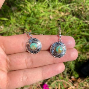 May include: A pair of silver-toned earrings with turquoise-colored stone centers. The earrings have a circular design with a decorative border. The earrings are displayed against a blurred green background.