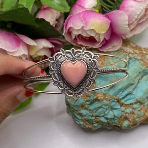 May include: A silver cuff bracelet featuring a central heart-shaped pink stone set within an ornate, scalloped silver frame. The band consists of two thin, smooth silver wires and one central twisted silver wire, creating a layered appearance.