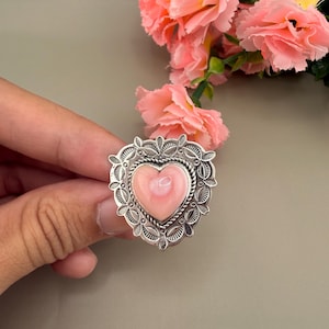 May include: A silver ring featuring a heart-shaped pink stone set in a decorative, filigree-style metal frame with a twisted rope border. The ring is positioned in front of a cluster of light pink carnation flowers with green stems.