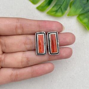 May include: A pair of rectangular stud earrings featuring a vibrant orange stone inlay set within a polished silver frame. Each earring has a distinct twisted rope border detail surrounding the central rectangular orange stone inlay.