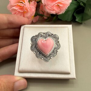 May include: A sterling silver ring featuring a smooth, heart-shaped pink conch shell cabochon set in the centre. The silver setting has a decorative scalloped edge with intricate etched line details surrounding the central heart stone.