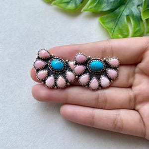 May include: A silver-toned brooch featuring two central oval turquoise cabochons surrounded by a cluster of smaller, teardrop-shaped pink stones. The metal setting has a dark, oxidized finish with beaded detailing around each individual stone.