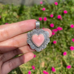 May include: A heart-shaped pendant featuring a smooth, light pink stone center set within a decorative, scalloped silver-toned metal frame. The metal border displays intricate etched line patterns and a small, textured bail at the top.