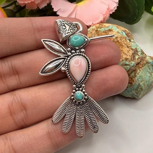 May include: A silver hummingbird pendant featuring a teardrop-shaped pink stone in the center, a small turquoise stone in the head, and another turquoise stone at the base of the tail. The wings and tail are crafted with detailed silver metalwork.