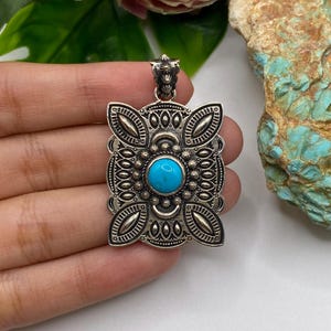 Puede incluir: Un colgante rectangular de plata con un intrincado diseño de metal estampado y un cabujón central de turquesa redonda. El colgante tiene un pasador decorativo en la parte superior y patrones simétricos que incluyen puntos.