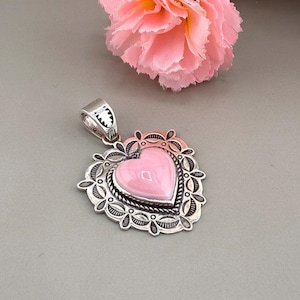 May include: A heart-shaped pendant featuring a smooth, light pink stone set in a decorative silver frame. The metal border displays intricate stamped patterns and a twisted rope detail surrounding the central stone, with a silver bail at the top.