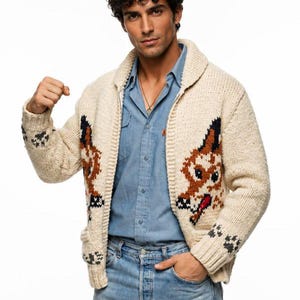 Può includere: Cardigan in maglia grossa color crema con un disegno a forma di muso di cane marrone e nero sul davanti e impronte di zampe sulle maniche. Il cardigan ha un collo a scialle e tasche. La persona indossa una camicia di jeans blu e jeans.