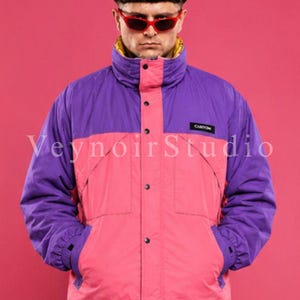 Op de afbeelding: Een color-blocked gewatteerd jack met een felpaars bovengedeelte en een felroze ondergedeelte. Het jack heeft een hoge kraag, een knoopsluiting aan de voorzijde en een kleine zwarte rechthoekige patch op de borst met de tekst CUSTOM.