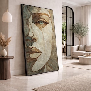 May include: Large framed artwork featuring a detailed, abstract portrait of a face. The artwork uses a combination of beige, brown, and teal tones. The portrait is leaning against a white wall in a well-lit room, suggesting a modern interior design theme.