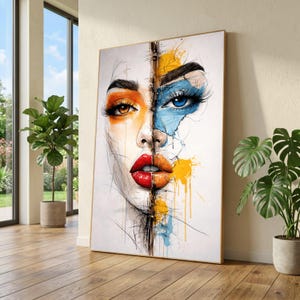 May include: A framed art print featuring a split-face portrait. One side is orange and the other is blue, with matching eye makeup. The lips are red and orange. The artwork has a watercolor and sketch style, with yellow and blue paint splatters.