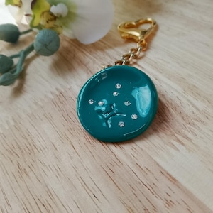 May include: Teal-colored round keychain with a gold-colored heart-shaped clasp. The keychain has a constellation design with small, sparkling crystals. The background includes a wooden surface and a white flower.