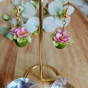 May include: Handmade earrings with miniature pink and yellow flower designs and green leaves. The earrings hang from gold-coloured hooks. The background includes a gold-coloured stand and white flowers.