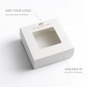 Puede incluir: Una caja de panadería cuadrada blanca con una ventana transparente. La caja tiene el texto "THE BAKER'S TABLE" y "FRESH & DELICIOUS" impreso en dorado. La caja también tiene el texto "ADD YOUR LOGO" y "AVAILABLE IN MULTIPLE FINISHES".