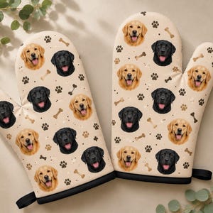 May include: A pair of beige oven mitts featuring a repeating pattern of golden retriever and black labrador faces, interspersed with small brown paw prints and bone shapes. Each mitt has a black trim at the wrist and a small black hanging loop.