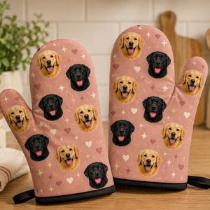 Könnte beinhalten: Ein Paar Ofenhandschuhe in Altrosa mit einem wiederkehrenden Muster aus Golden Retriever- und schwarzen Labrador-Gesichtern, durchsetzt mit kleinen weißen Herzen und vierzackigen Sternen, mit schwarzem Saum am Handgelenk.
