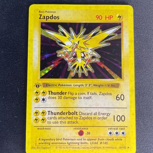 Puede incluir: Una carta coleccionable Zapdos amarilla con una ilustración holográfica del Pokémon pájaro legendario. La carta muestra 90 HP, los ataques Trueno y Rayo, un sello de primera edición y una altura de 1,6 m con un peso de 52,6 kg.