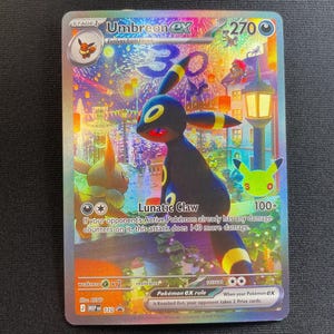 May include: A holographic Umbreon ex trading card featuring a dark blue creature with yellow rings, standing in a vibrant town square at night with fireworks and lanterns. The card displays 270 HP, the Lunatic Claw attack, and a 30th anniversary logo.