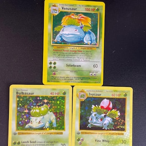 Könnte beinhalten: Drei Sammelkarten mit den Pokémon-Charakteren Bisaflor, Bisasam und Bisaknosp. Jede Karte hat einen gelben Rand, grüne Textfelder und zeigt den Namen des Charakters, die KP, Angriffsfähigkeiten sowie beschreibenden Text auf Englisch.