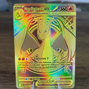 May include: A gold-colored Mega Charizard Y EX trading card featuring a holographic rainbow finish. The card displays the character illustration, 360 HP, the Explosion Y attack text, and the card number 294/217. It is displayed on a clear plastic stand.