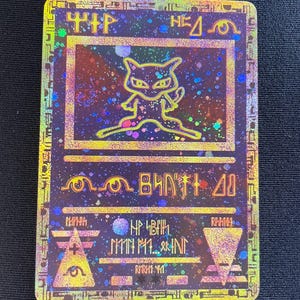 May include: A rectangular holographic trading card featuring a stylised gold outline of the creature Mew centred in a frame. The card has a dark purple background with scattered colourful glitter effects and ancient-style symbols and text.