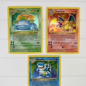 May include: Three collectible trading cards featuring Venusaur, Charizard, and Blastoise. Each card displays character artwork, 100 to 120 HP, attack statistics, and descriptive text. The cards are arranged in a triangular formation on a white surface.