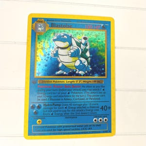 May include: A holographic Blastoise trading card with a yellow border and blue background. The card features the character Blastoise, 100 HP, the text Shellfish Pokémon, Length: 5' 3", Weight: 189 lbs., and the attacks Rain Dance and Hydro Pump.