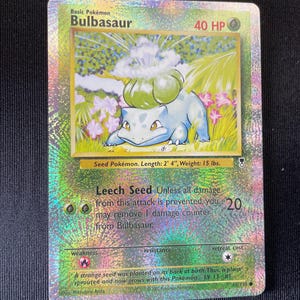 May include: A holographic Bulbasaur trading card featuring the character with a green bulb on its back. The card displays 40 HP, the Leech Seed attack, and text including Seed Pokémon, Length: 0.71 m, Weight: 6.8 kg, and a descriptive Pokédex entry.