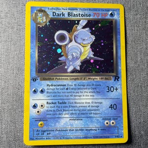 May include: A holographic Dark Blastoise trading card with a yellow border. The card features the character Dark Blastoise, 70 HP, and the attacks Hydrocannon and Rocket Tackle. It includes the 1st Edition stamp, level 28, and number 3/82.