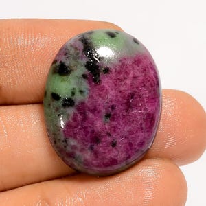 May include: An oval-shaped gemstone with a polished surface. The stone features a vibrant color palette, with a deep magenta hue dominating the lower half and a light green shade on the upper half, speckled with black inclusions.
