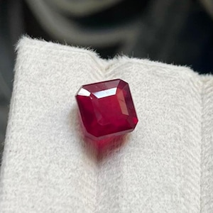 Op de afbeelding: Een vierkant geslepen, dieprode edelsteen met gefacetteerde oppervlakken die het licht reflecteren. De steen heeft een levendige, verzadigde karmozijnrode kleur en een gepolijste afwerking, met een heldere, geometrische vorm.