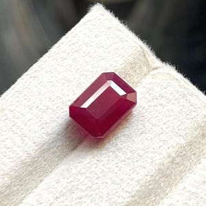 Op de afbeelding: Een dieprode, rechthoekige edelsteen met een emerald slijpvorm, voorzien van gefacetteerde oppervlakken die het licht reflecteren. De steen vertoont een rijke, verzadigde karmozijnrode kleur op een getextureerd, gebroken wit oppervlak.