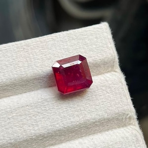 Op de afbeelding: Een dieprode, vierkant geslepen edelsteen met een gefacetteerd oppervlak dat licht reflecteert. De steen heeft scherpe, strakke randen en een levendige, verzadigde karmozijnrode kleur, met een klassieke geometrische vorm en gepolijste facetten.