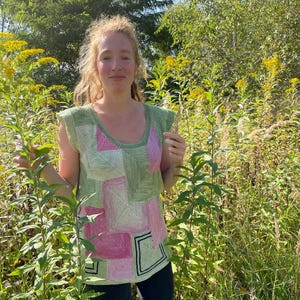 Könnte beinhalten: Ein ärmelloses Oberteil im Patchwork-Stil in Grün- und Rosatönen. Das Oberteil hat ein geometrisches Design mit quadratischen und rechteckigen Mustern. Der Halsausschnitt und die Armausschnitte sind grün eingefasst. Die Person steht in einem Feld.